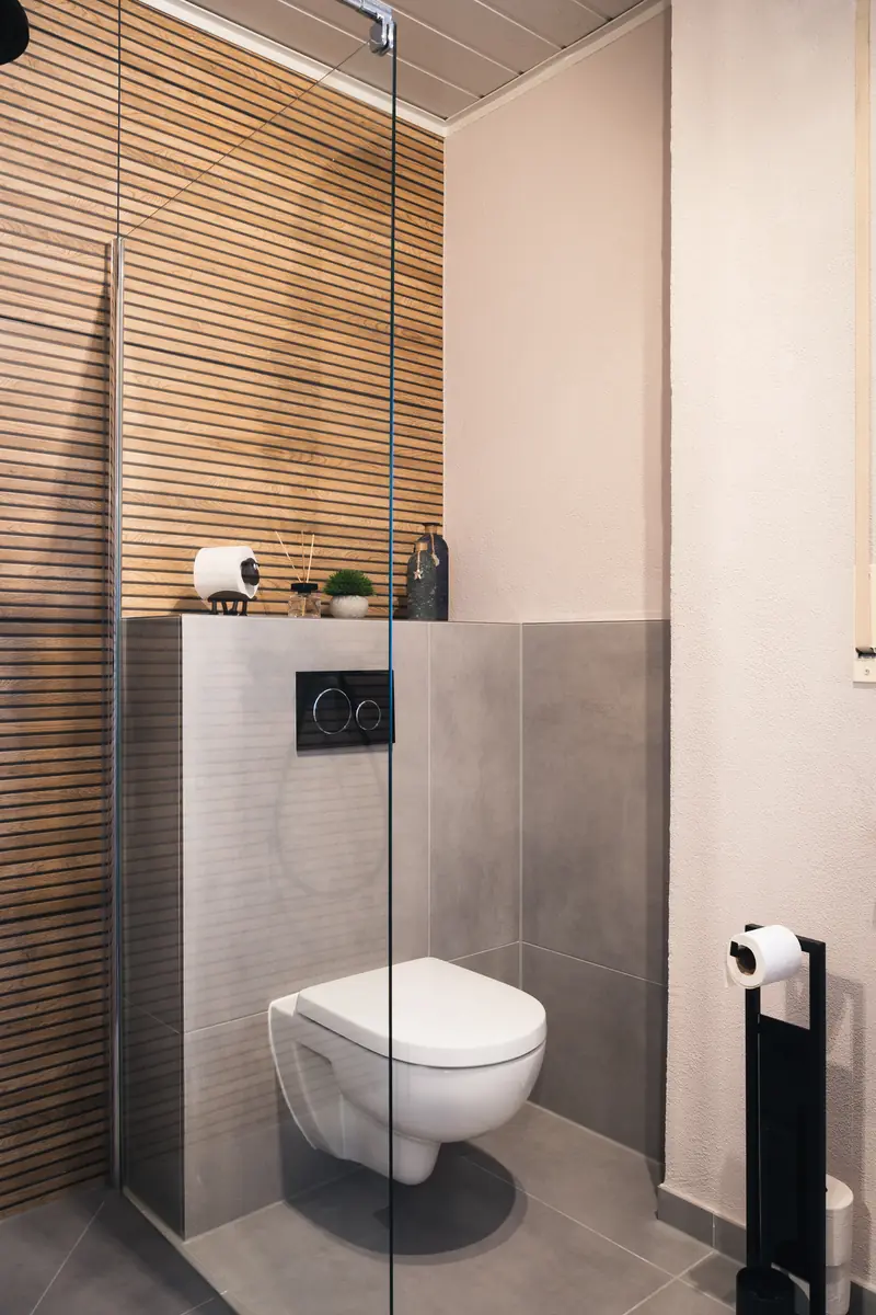 WC, durch eine Glaswand von der Dusche getrennt, mit Wandverkleidung in Holzoptik